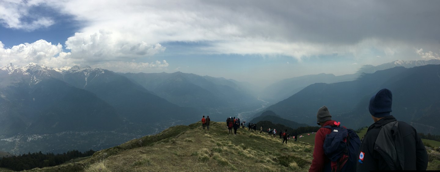 Trekkers walk along a grassy ridge towards snow-capped mountains.